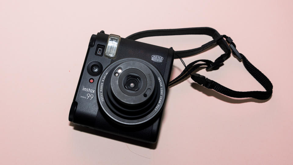 A photograph of the Fujifilm Instax mini 99 in black, set against a pink background.