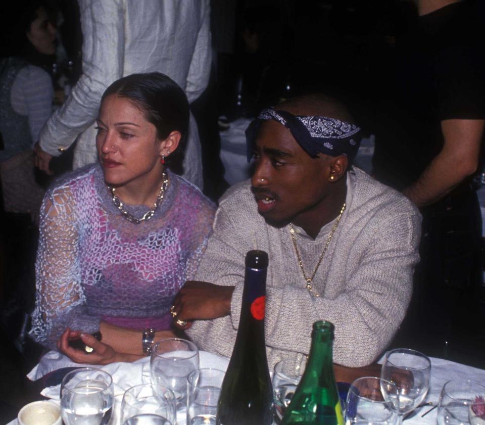 An old photo of Madonna and Tupac sitting at a table that's covered in glasses and wine bottles, in conversation with someone to their right