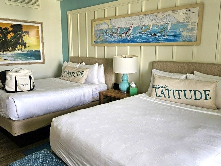 A hotel room with two queen beds, ocean- and beach-themed paintings on the yellow walls, and a blue bench at the foot of the bed on the right.