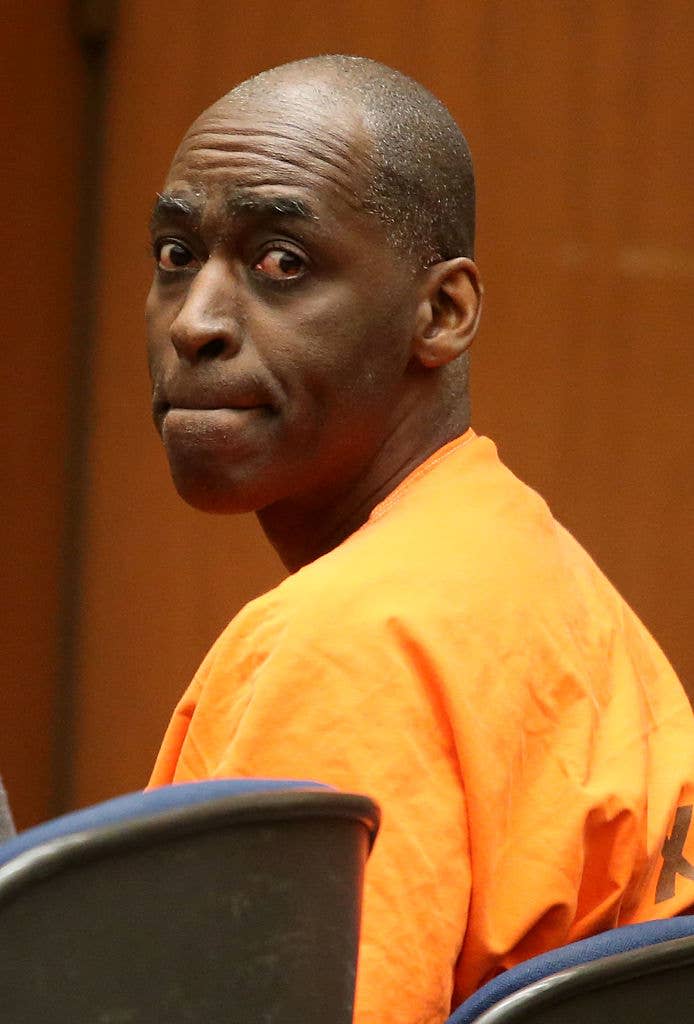 A bald man wearing an orange shirt is seated in a courtroom, looking over his shoulder with a serious expression