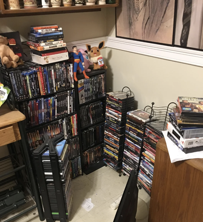 A cluttered corner room with numerous stacked DVDs and Blu-rays, superhero and Pokémon toys on the shelves, and various decorative items