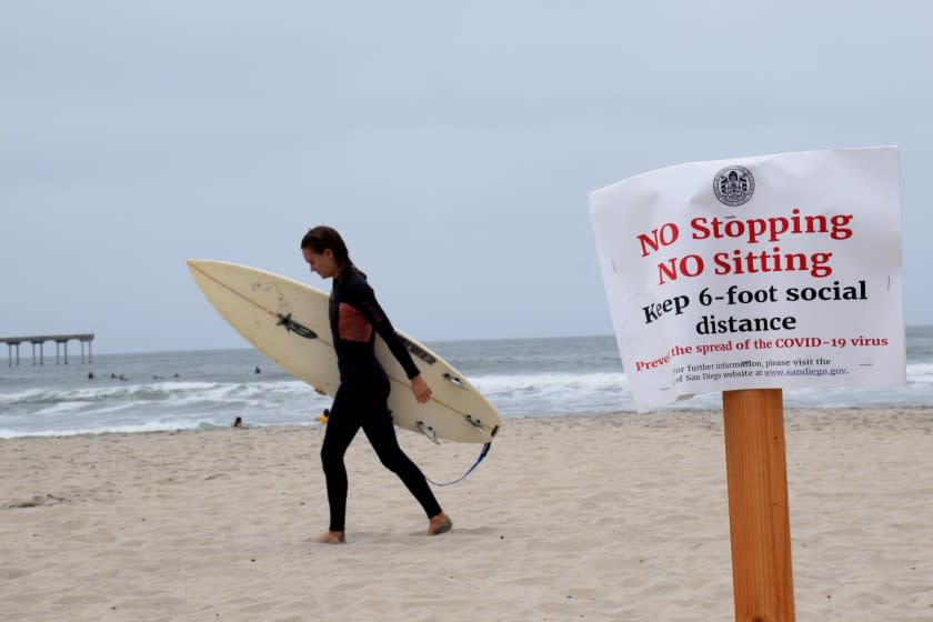 A Memorial Day surfer walks by a sign warning beach goers not to stop or sit on the beach due to COVID-19 social distancing guidelines in Ocean Beach on March 25, 2020.