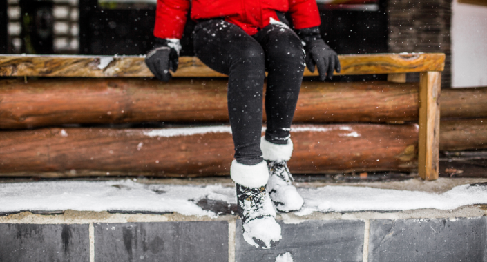 woman in red jacket, black jeans, and snow boots with snow on them sitting outside