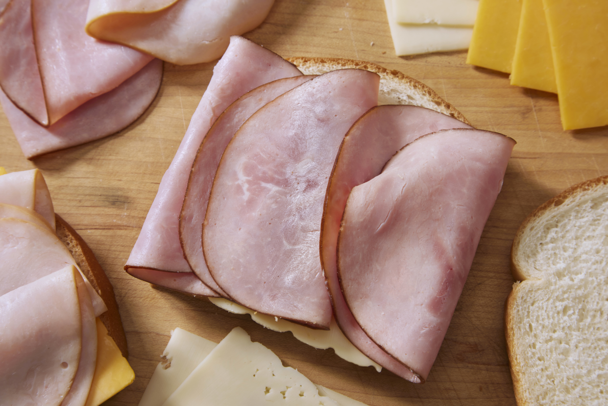 Focus on one half of a monte cristo sandwich, with black forest ham, smoked turkey breast, cheddar, and swiss cheese on a wooden cutting board, surrounded by ingredients