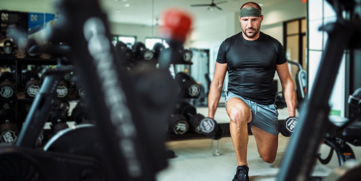 a fit and healthy middle aged caucasian man does a lunge while holding two weighted dumbbells he is concentrating on working out in an indoor gym