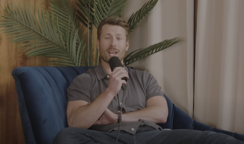 Glen Powell sits on a blue chair, holding a microphone, in a relaxed pose during an interview. Palm leaves are visible in the background