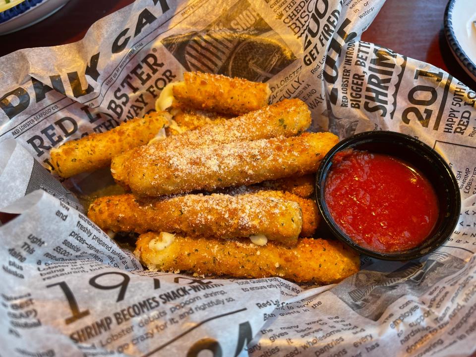 Mozzarella sticks dusted with cheese next to a black cup filled with a marinara sauce and newspaper-print paper