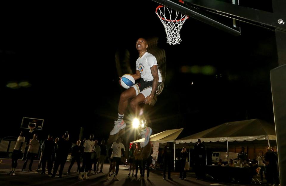 Tyler Currie jumps in the air with a basketball near a hoop while several people watch at night near some tents.