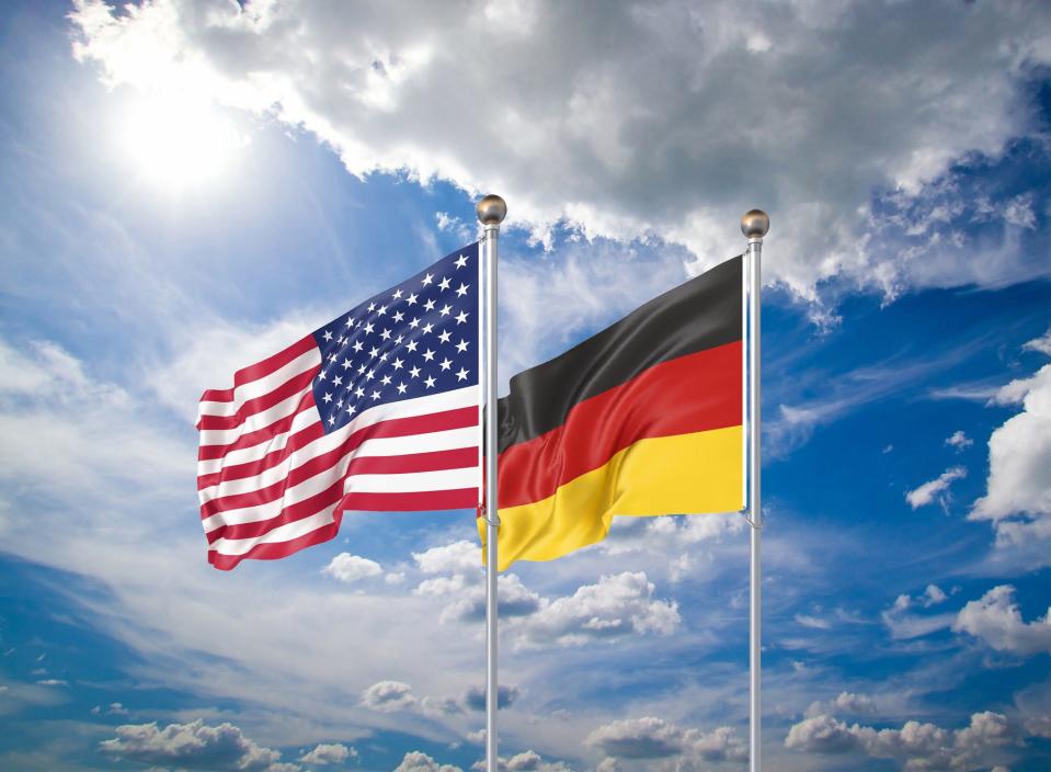 The United States flag and German flag fly in the wind against a background with blue skies and clouds.