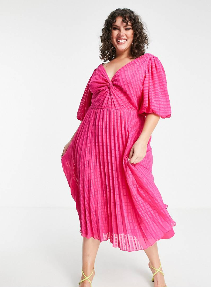 A woman with dark shoulder length curly hair wears an ASOS DESIGN Curve twist front pleated midi skater dress in dobby in hot pink - $90.00 Photo: ASOS