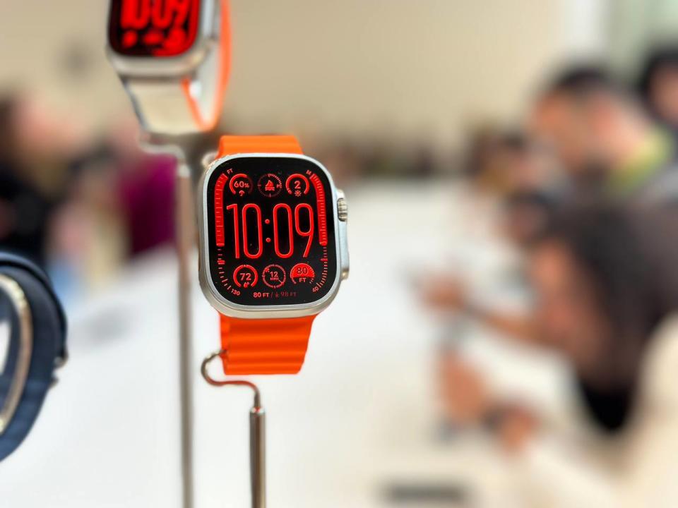 The Apple Watch Ultra 2 with an orange strap, hanging on a stand. Its screen shows the new modular ultra face in night mode, with red wording and graphics.