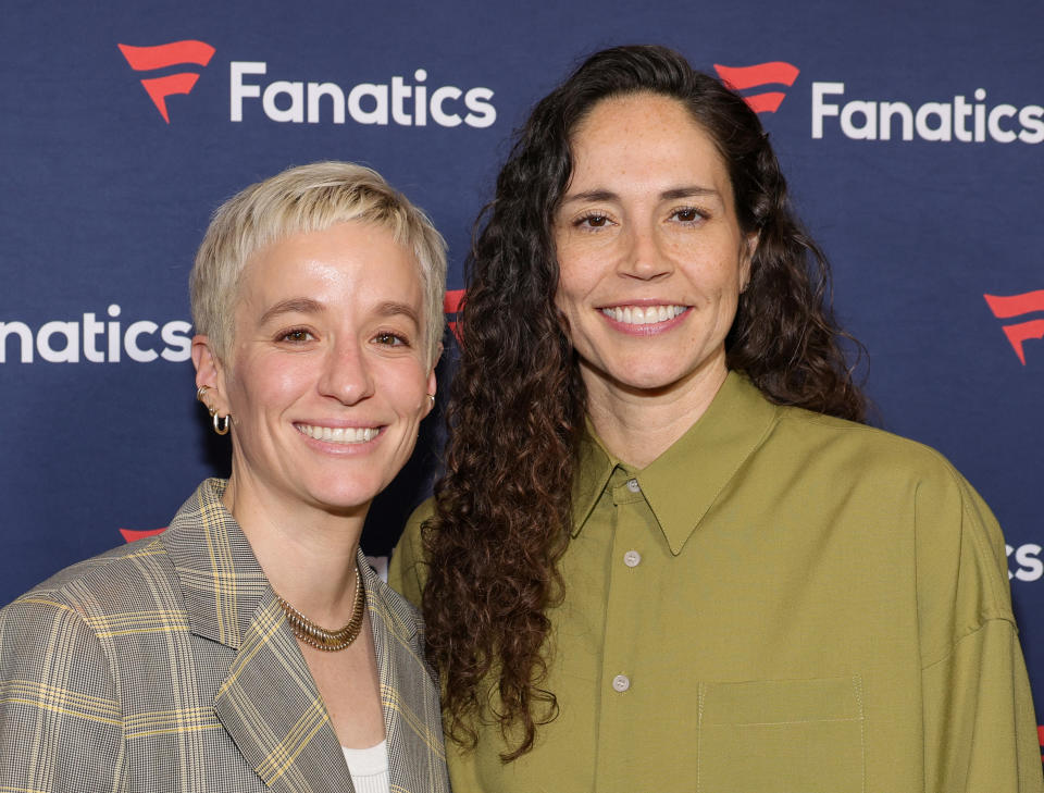 Megan Rapinoe and Sue Bird at a Fanatics event, smiling, with Megan in a plaid blazer and Sue in a green button-up shirt