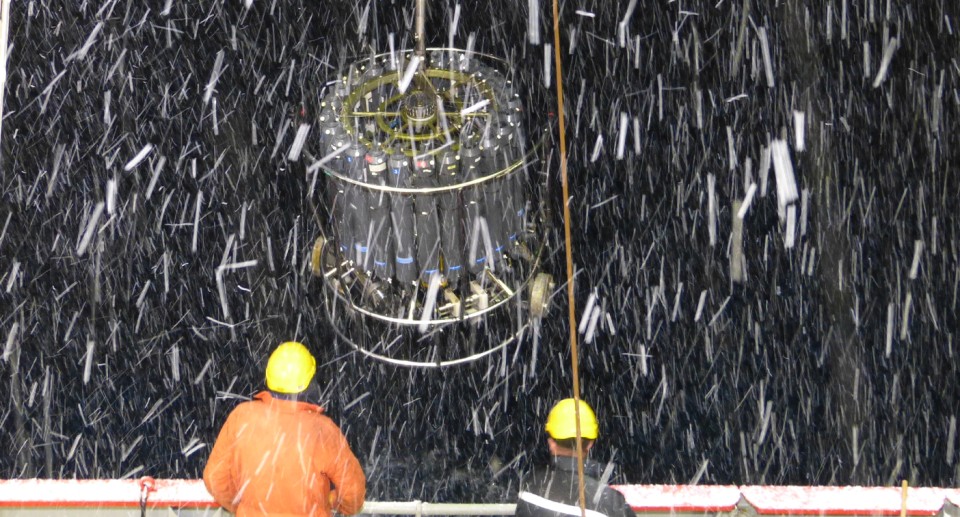 In the foreground we see the backs of two men in helmets. It is snowing. A Conductivity-Temperature-Depth (CTD) is being hauled in. It is dark.