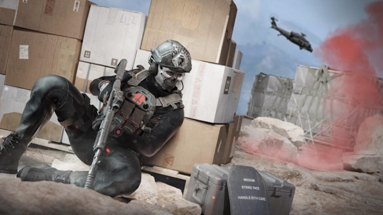  A soldier crouching behind cover as a helicopter flies over red smoke markers in the distance. 