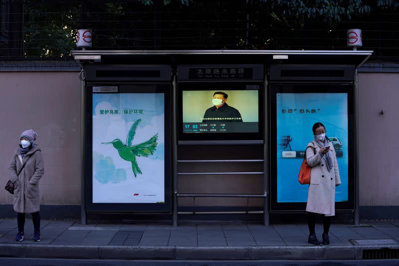 Women wearing masks stand in front of a TV screen showing news of Chinese President Xi Jinping at a bus station in Shanghai