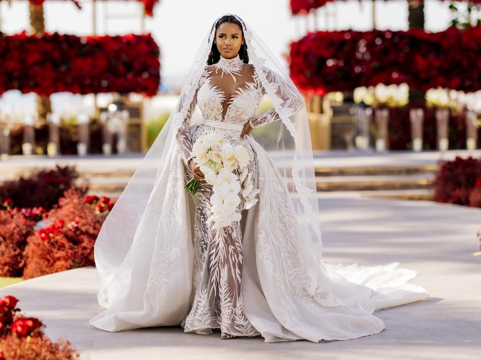 A bride stands in her wedding dress in front of elaborate arrangements of red flowers.