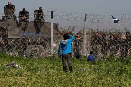 A boy stands next to a border fence on the Greek side of the border as Macedonian police stand guard on the Macedonian side, close to a makeshift camp for refugees and migrants near the Greek village of Idomeni, April 12, 2016. Picture taken from the Greek side of the border. REUTERS/Alexandros Avramidis