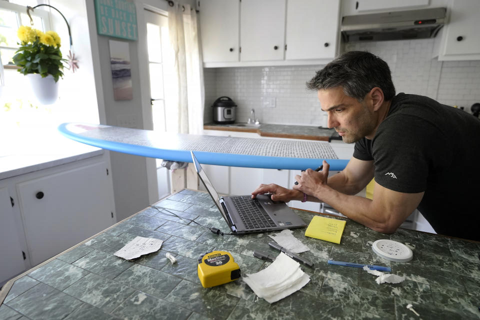 Dan Fischer uses a computer while placing the names of lost loved ones on one of his surfboards at his home, in Newport, R.I., Wednesday, May 25, 2022. Fischer, 42, created the One Last Wave Project in January 2022 to use the healing power of the ocean to help families coping with a loss, as it helped him following the death of his father. Fischer places the names onto his surfboards, then takes the surfboards out into the ocean as a way to memorialize the loved ones in a place that was meaningful to them. (AP Photo/Steven Senne)