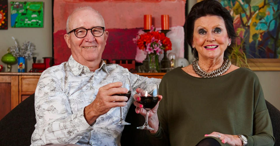 Gogglebox Couple Mick and Di hold up glasses of wine while smiling at the camera. Mick is wearing a white shirt with a simple print, Di is wearing an olive green jumper