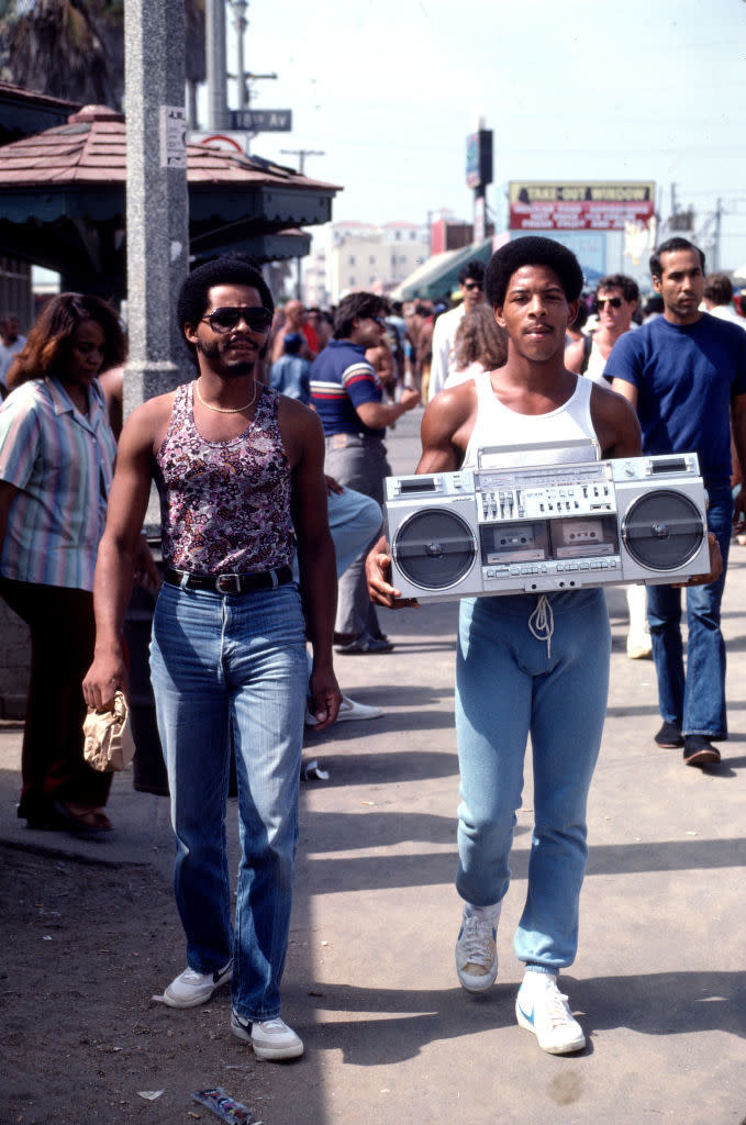 Two men in 1980s casual attire walking outside; one carries a boombox. People in the background. Names unknown