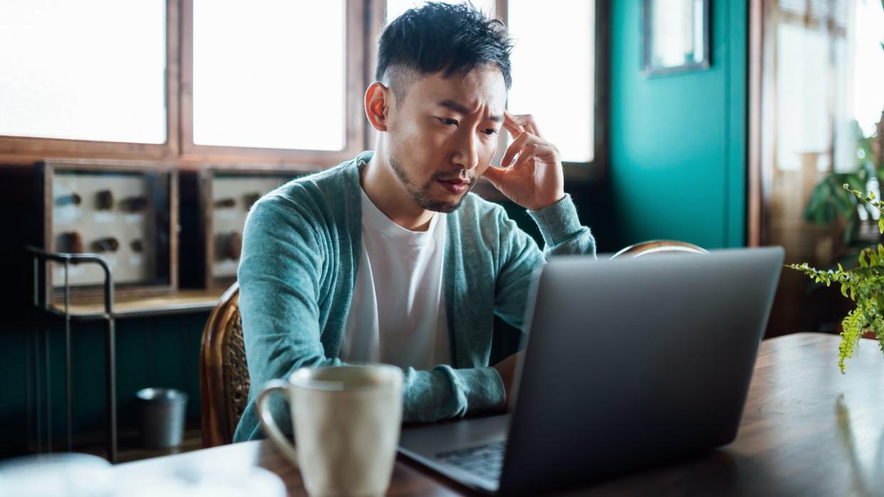 worried young asian man with his hand on head, using laptop computer at home, looking concerned and stressed out