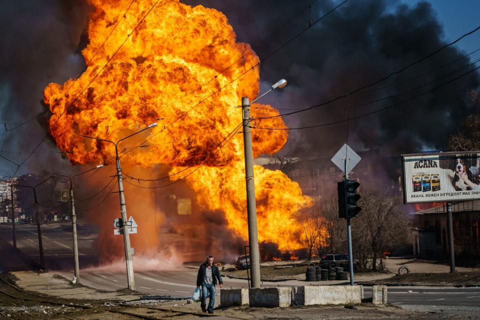 A man walks away from a burning building with bright orange flames filling the sky behind him