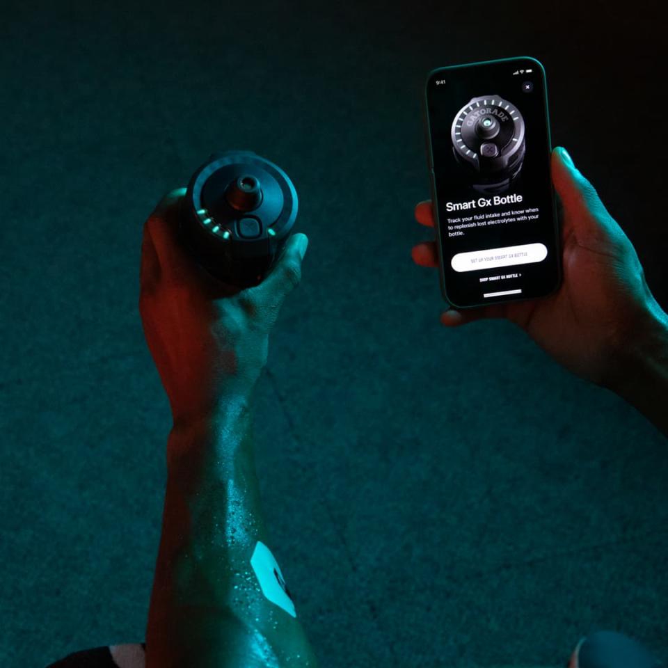 A person holds the Gatorade Smart Gx Bottle, which has a cap with lights around the edge. In their other hand, they're holding a smartphone that displays the Gx App with text reading Smart Gx Bottle.