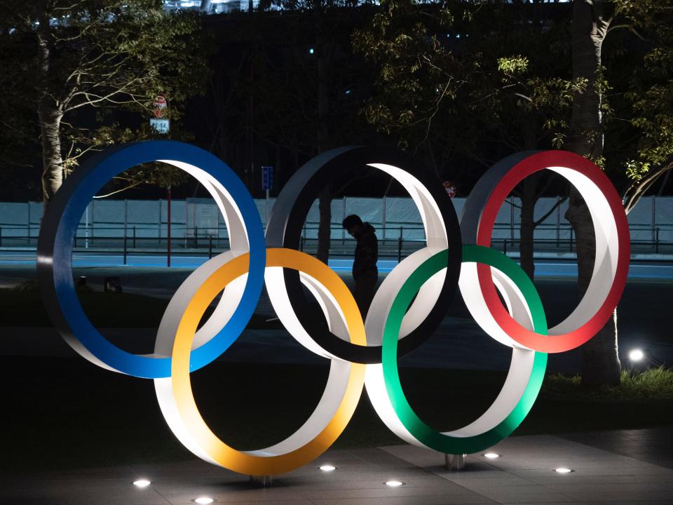 A man is seen through the Olympic rings in front of the New National Stadium in Tokyo, Tuesday, March 24, 2020. IOC President Thomas Bach has agreed "100%" to a proposal of postponing the Tokyo Olympics for about one year until 2021 because of the coronavirus outbreak, Japanese Prime Minister Shinzo Abe said Tuesday. (AP Photo/Jae C. Hong)