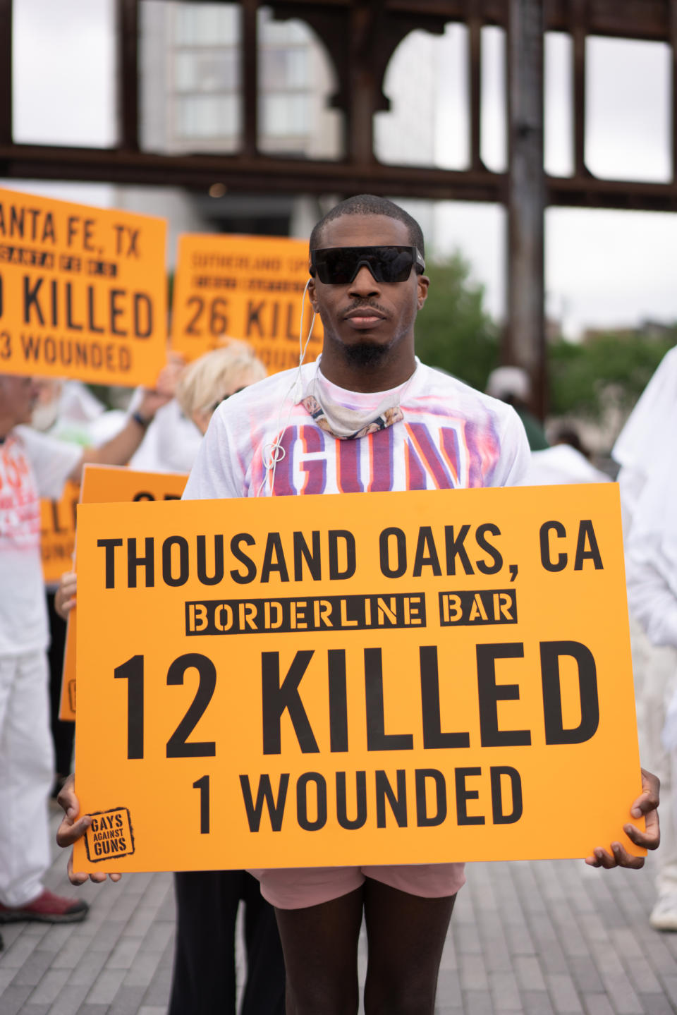  A protester wearing sunglasses with a face mask dangling around his neck stands outside distanced from other protesters, holding a sign reading: Thousand Oaks, CA, Borderline Bar, 12 killed, 1 wounded, Gays Against Guns.