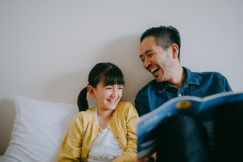 A man and a young girl share a joyful moment while reading a book together on a couch. The girl is wearing a yellow sweater