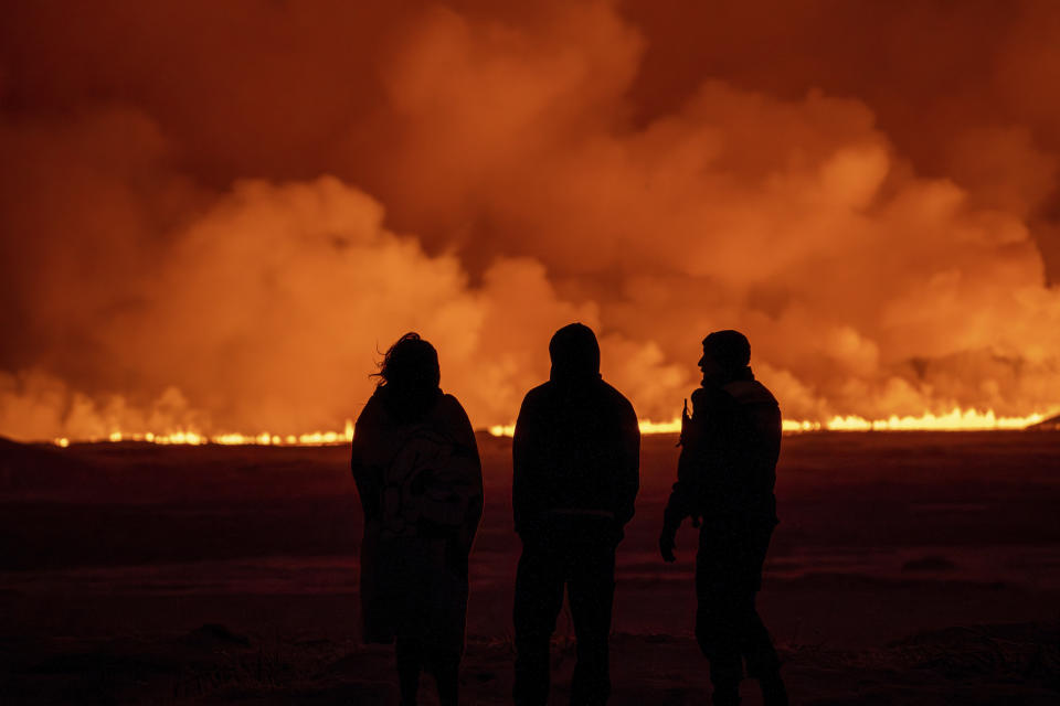 Gente observando el cielo nocturno iluminado por la erupción de un volcán en Grindavik, en la península islandesa de Reykjanes, el lunes 18 de diciembre de 2023. (AP Foto/Marco Di Marco)