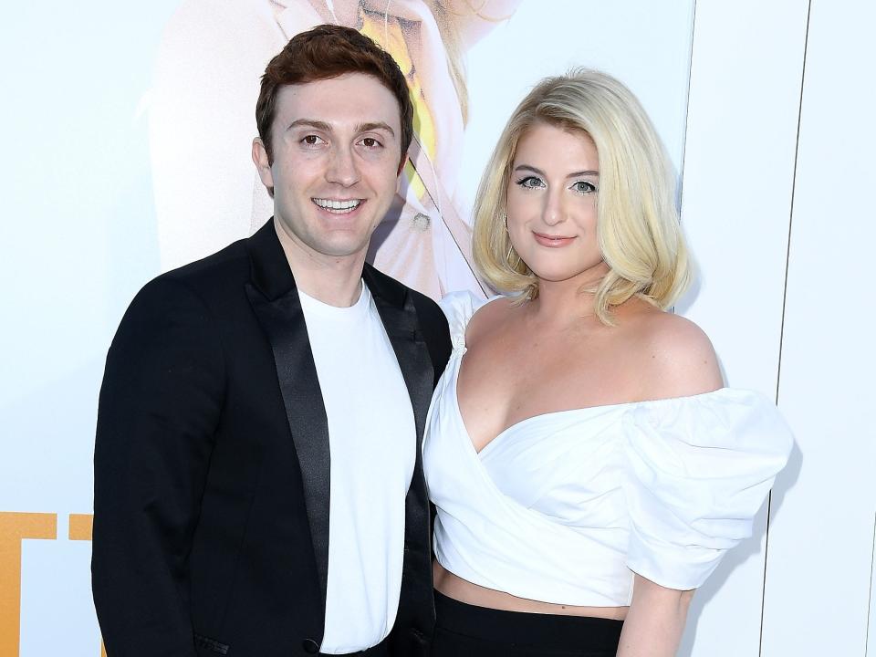 daryl sabara and meghan trainor at the premiere of i feel pretty, posing together on the red carpet. sabara is wearing an unbottoned black suit, while trainor is wearing black pants, an off the shoulder white crop top, and loosely curled hair.