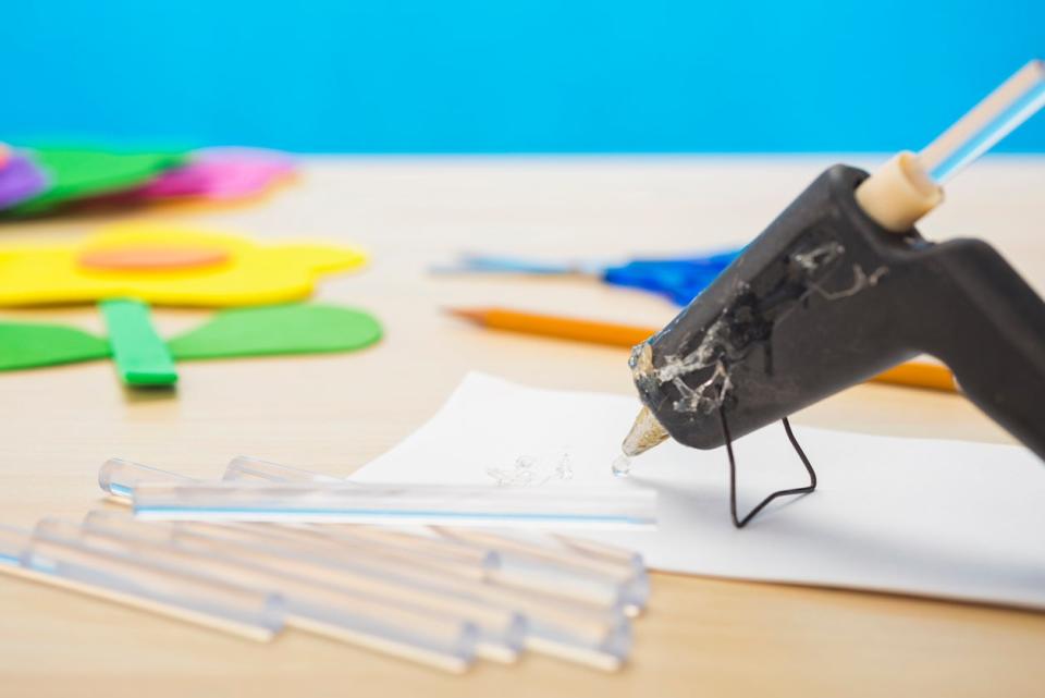 A mini hot glue gun with a blob of clear glue oozing out of the tip onto a scrap piece of white paper, with scissors and a pencil, along with a yellow flower created from foam sheets on a light colored wood grain table and a blue wall in the background.