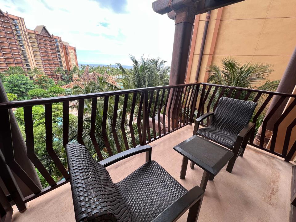 A hotel room balcony with chairs and a table and views of lush trees, pools, and the ocean in the distance.