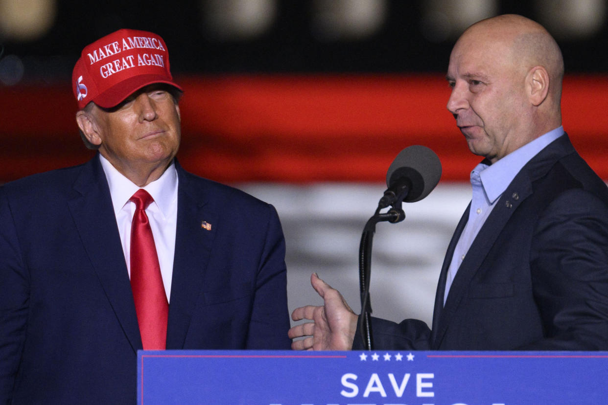 President Donald Trump, in red baseball cap saying Make America Great Again, looks on with a half-smile, as Doug Mastriano takes the microphone.