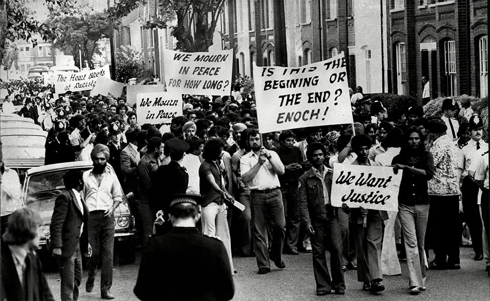 Black & white still of protests, with a crowd of people marching and holding signs.