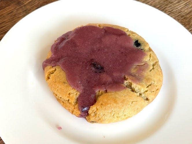 A cookie with blueberries inside and a blueberry-buttermilk glaze on top. The cookie is on a white plate.