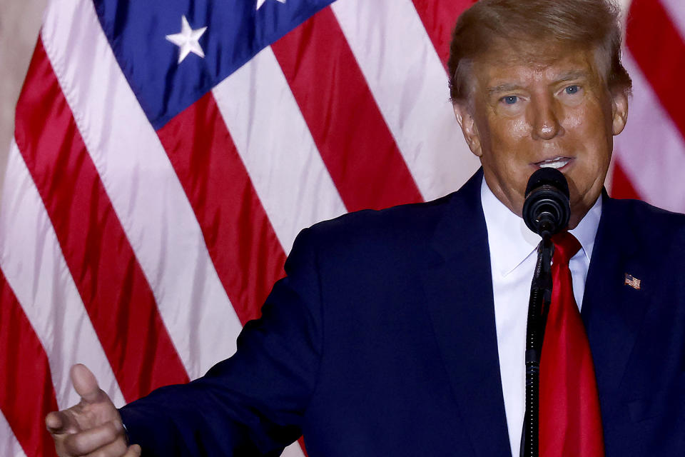 Donald Trump, in navy suit and red tie, emphasizes his point at the microphone, with an American flag behind him.