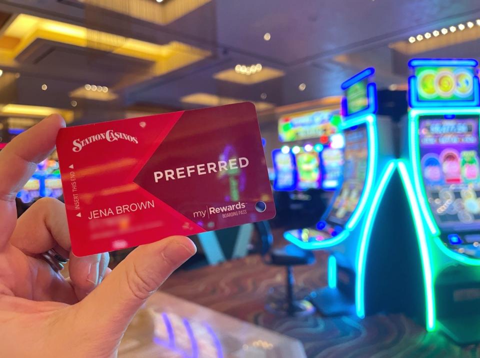 A hand holding up a Station Casinos rewards card while standing in front of a row of casino machines in a brightly-lit room.