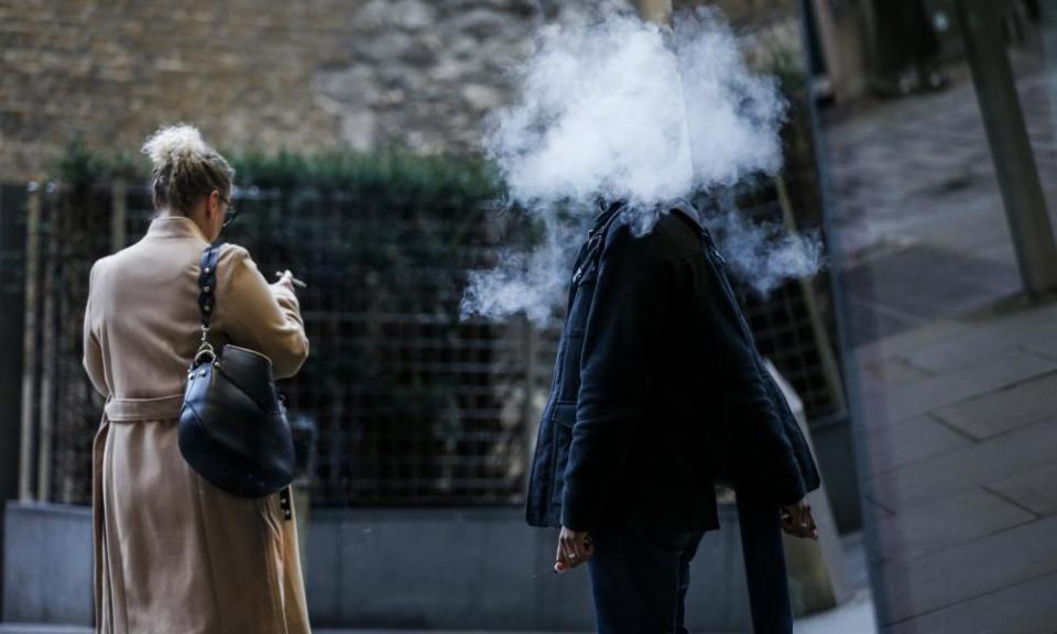 A pedestrian stands surrounded in a cloud of vapour after exhaling from a vape device as a pedestrian smoking a cigarette passes in London