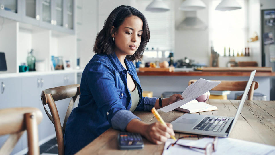 Shot of a young woman using a laptop and  going through paperwork while working from home.