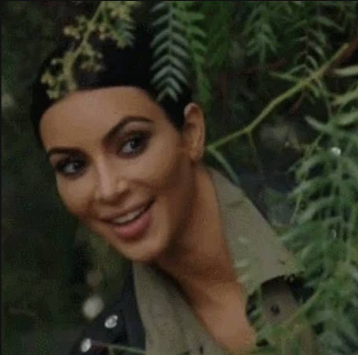 Kim Kardashian peers through tree branches, smiling. She wears a jacket with a lapel shirt underneath