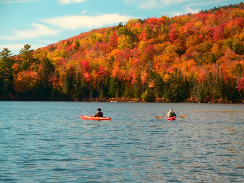 Two people paddle kayaks on Kettle Pond in Groton, Vermont with a background of a forest in autumnal colors of orange, red, and yellow