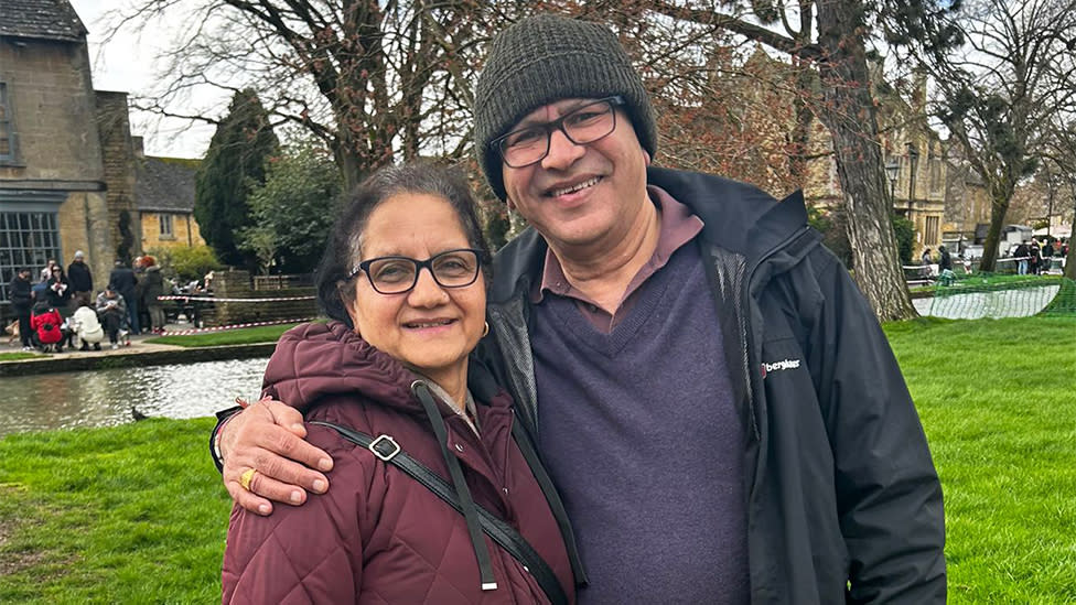 Meena Kumari's parents, smiling at the camera. Her mum is wearing glasses and a purple jacket, with her dad wearing a dark jacket, purple jumper. There are trees in the background and green grass.