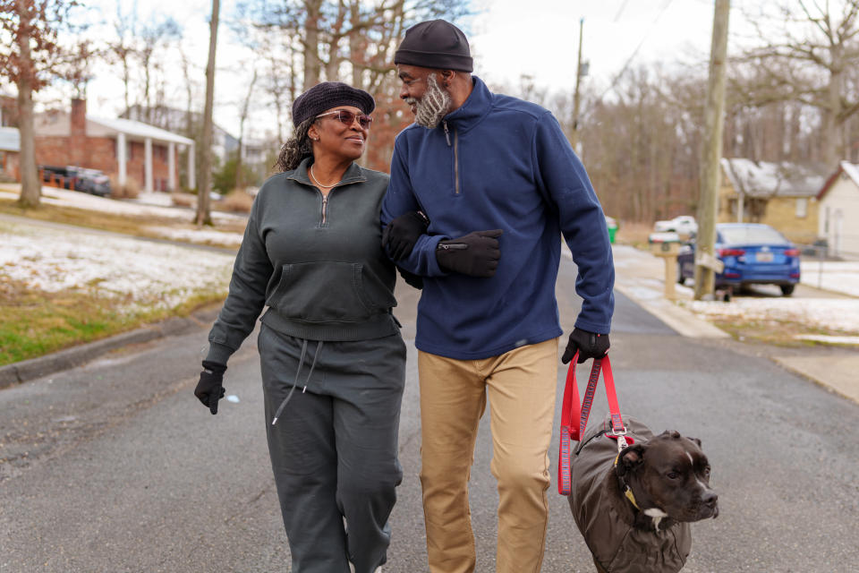 An older couple walks arm-in-arm with their dog on a neighborhood street, smiling and enjoying each other's company