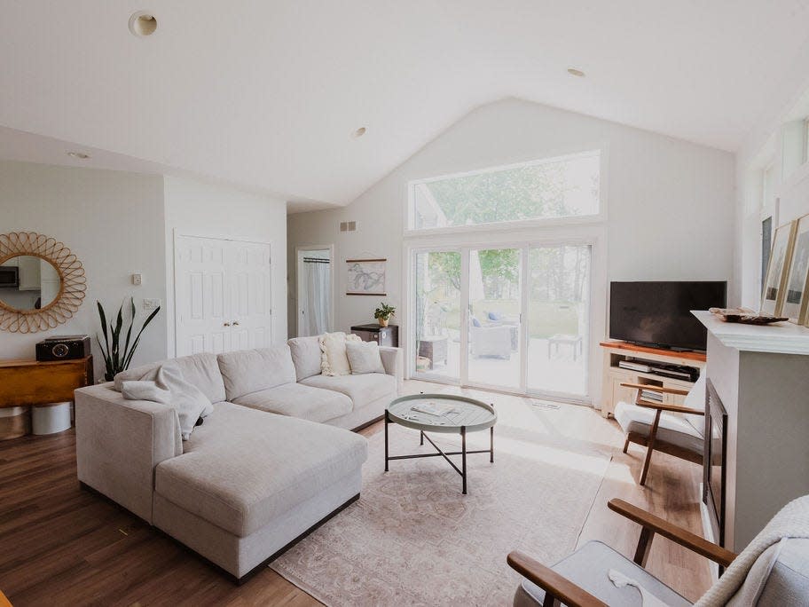 A white living toom with wood furniture, a white couch and rud, and windows in the back leading to a porch outside.