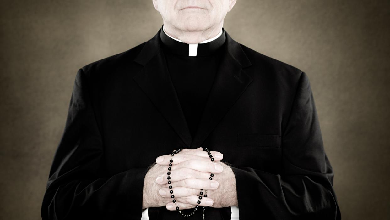  A faceless shot of a priest with his hands clasped, holding a rosary. 