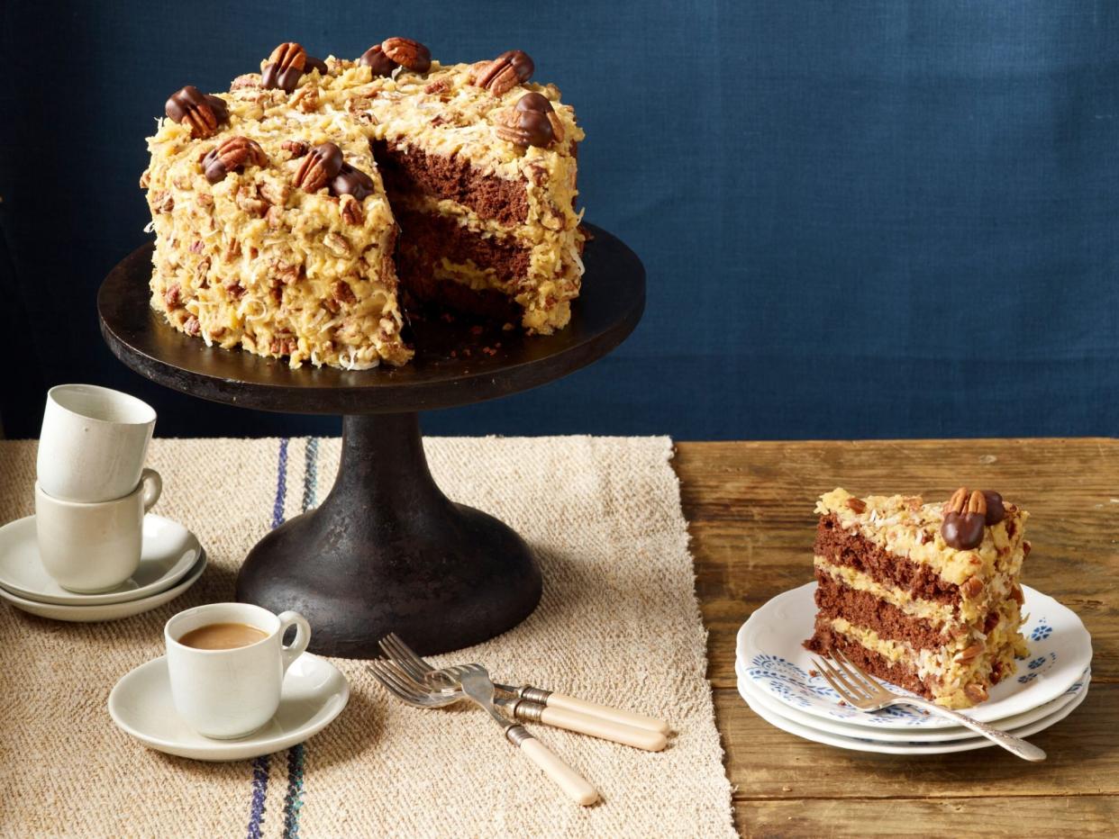 Close-up, Mama's German Chocolate Cake with Coconut-Pecan Frosting garnished with chocolate-dipped toasted pecan halves on footed cake stand beside cup of coffee and slice of cake on stack of plates.