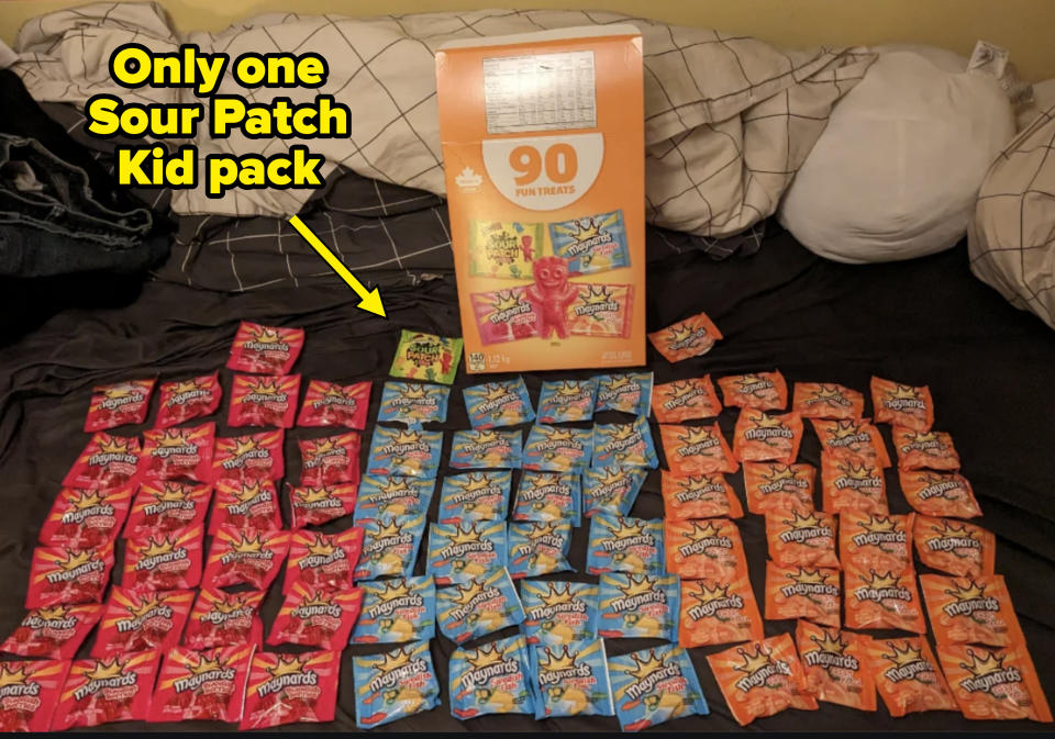 A large box labeled "90 Fun Treats" is surrounded by sorted piles of Sour Patch Kids candy packs on a bed