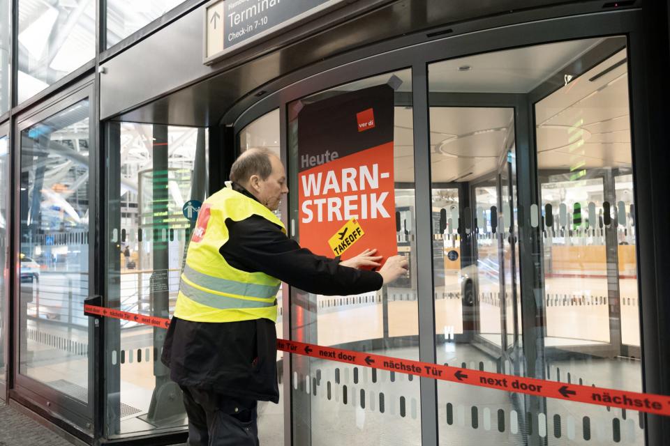 Ein Verdi-Vertreter hängt ein Plakat mit der Aufschrift "Warnstreik" an einer geschlossenen Terminaldrehtür am Hamburger Flughafen auf. - Copyright: picture alliance/dpa | Bodo Marks